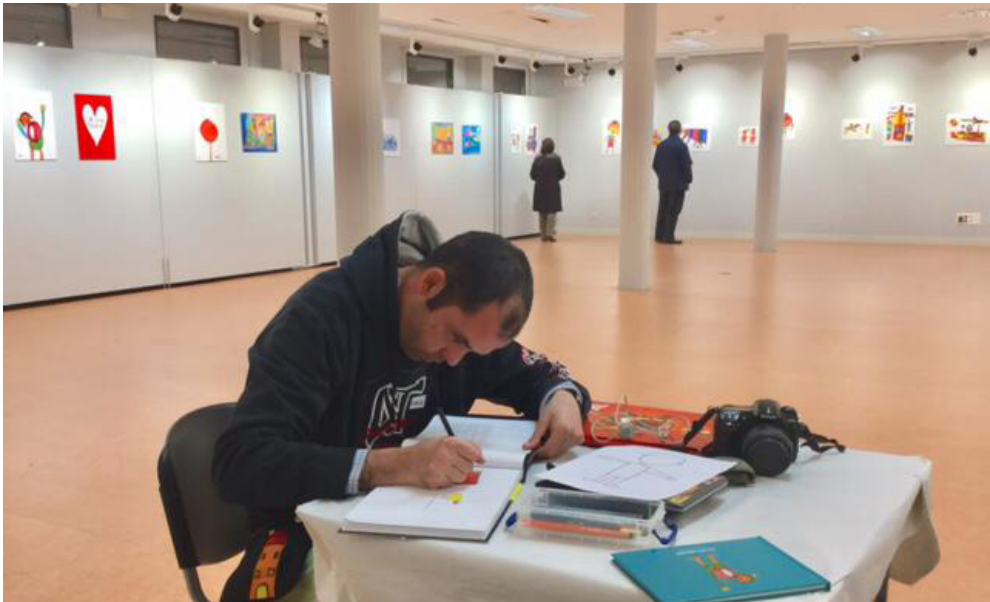 Raúl Águirre dibujando en una mesa en un espacio que expone sus obras